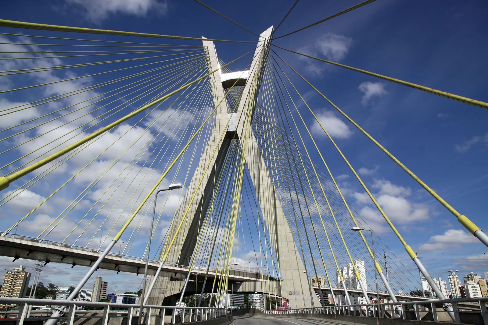 The Octávio Frias de Oliveira cable-stayed bridge from below, its white X-shaped pylon and fans of steel cables against a blue sky with the city behind