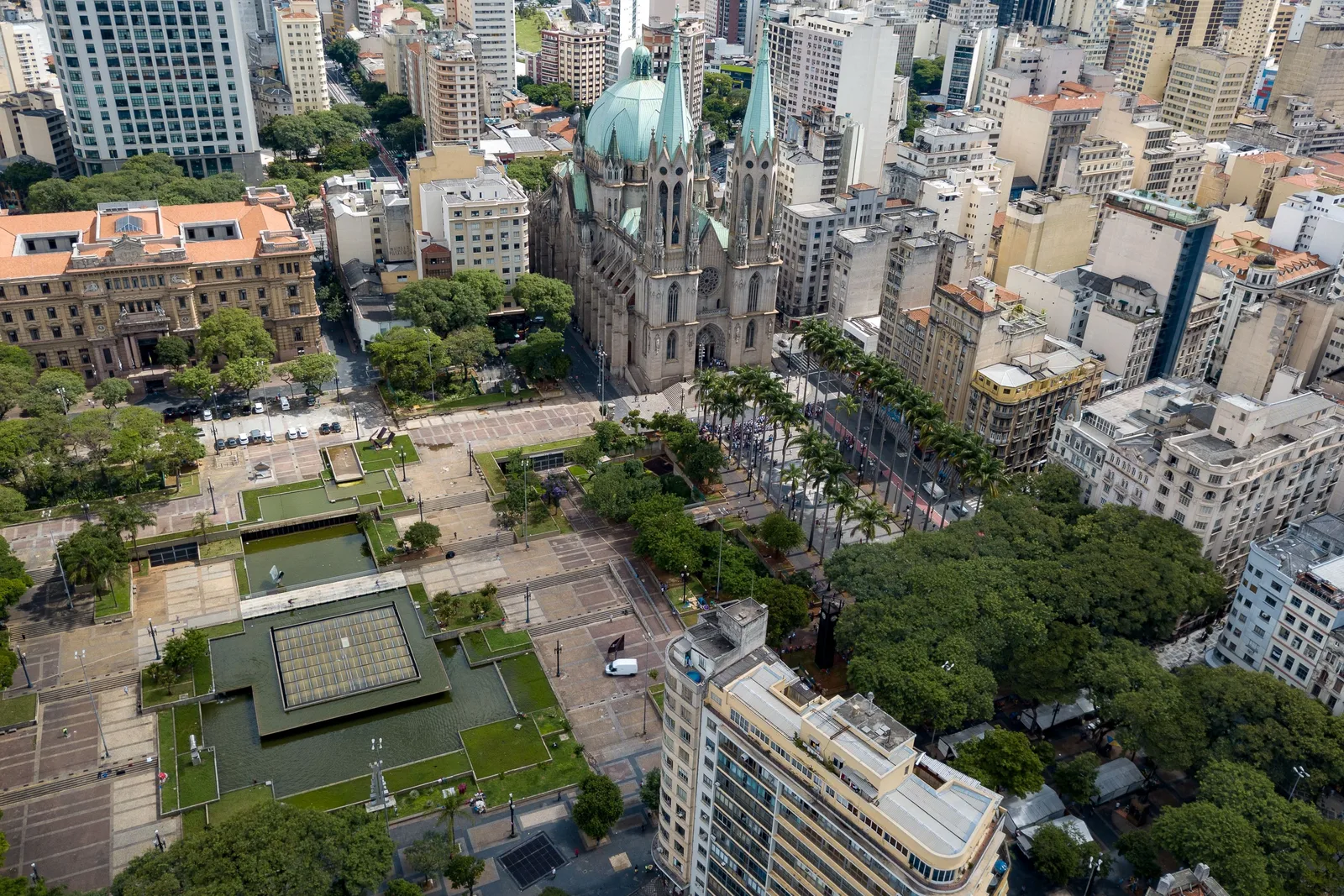 Central São Paulo from the air: the Praça da Sé filling the middle of the frame with its long rectangular pools and pale paving, a line of palms across it, the green dome and twin spires of the cathedral rising directly behind, and dense office blocks packed to every edge