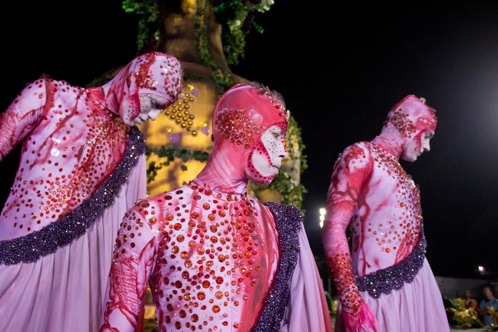 Three dancers of a São Paulo samba school on the Anhembi runway at night in tall pink and magenta beaded costumes, lit against the dark