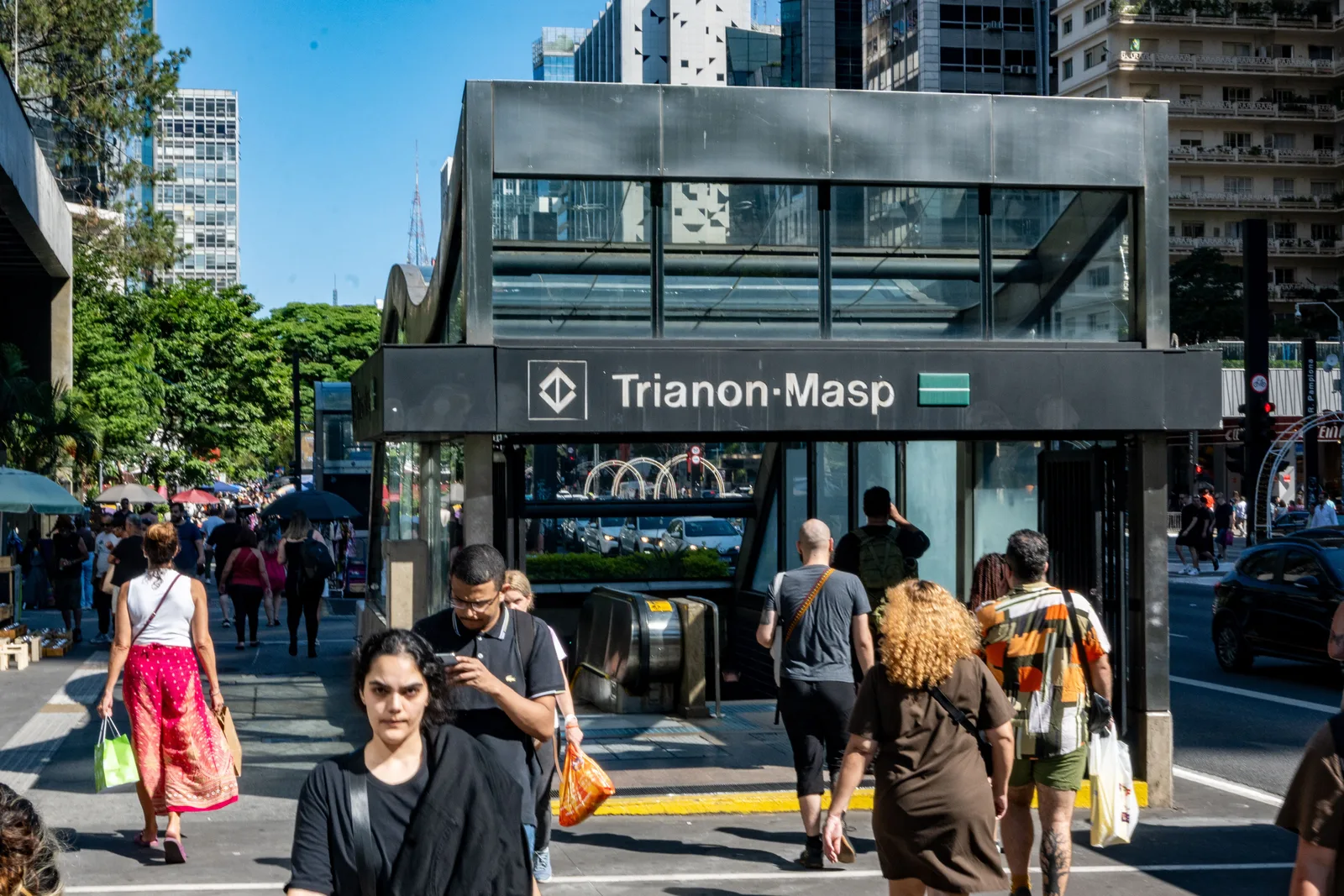 The glass entrance to Trianon-MASP metro station on Avenida Paulista, pedestrians crossing in front of it with shopping bags on a bright weekday afternoon