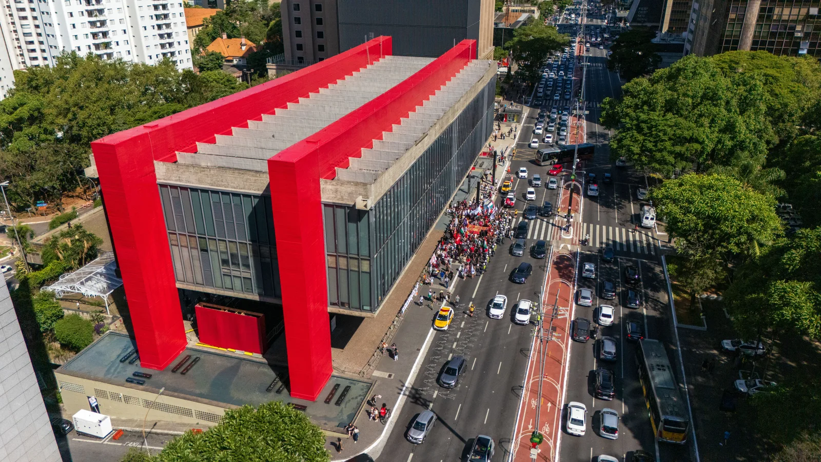 MASP from the air on Avenida Paulista: the museum's glass box slung between two bright red concrete beams over an open plaza, with traffic and a red bike lane running past and trees crowding both sides of the avenue