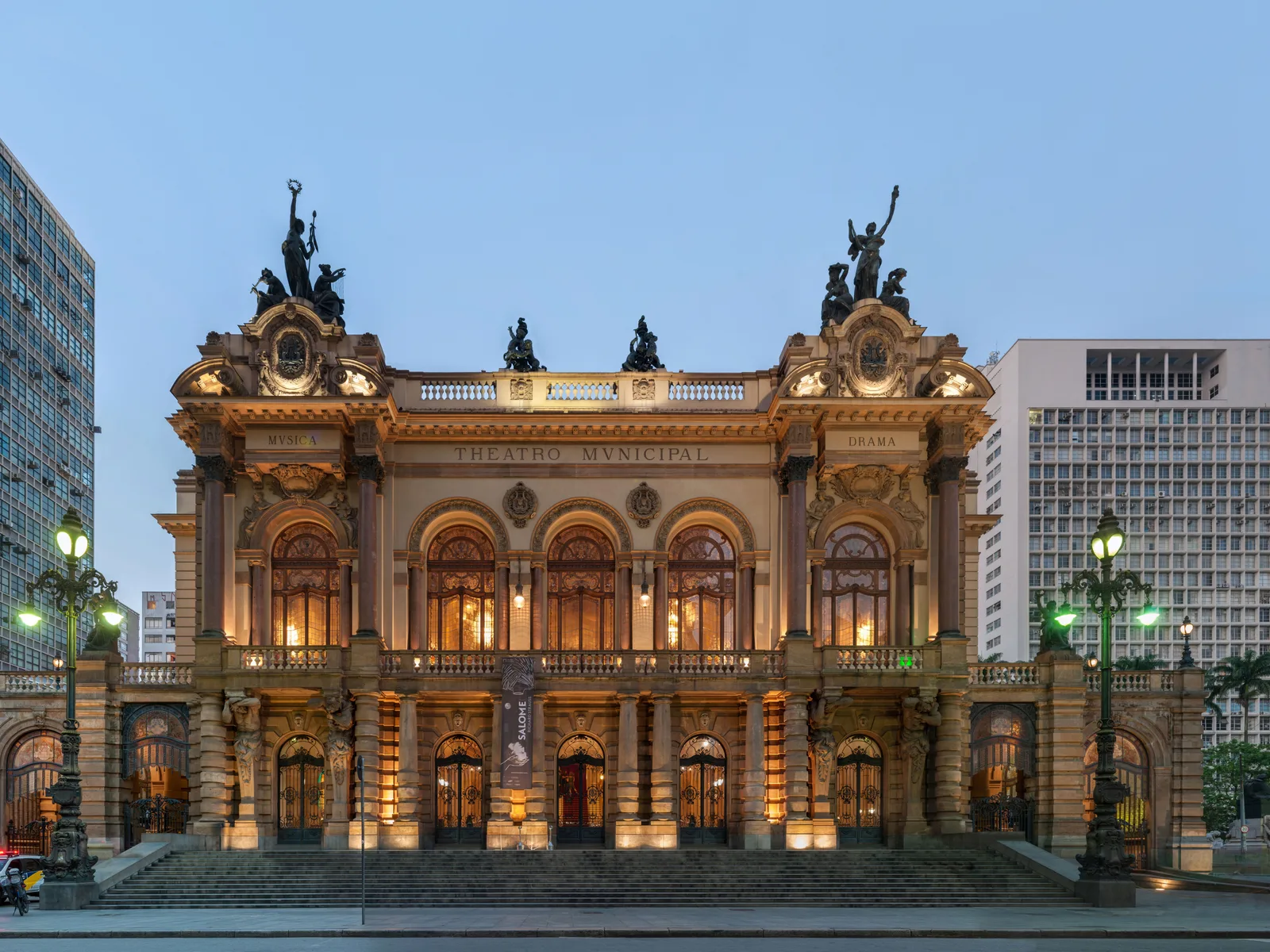 The Theatro Municipal floodlit at dusk: a Belle Époque opera house in pink stone and green bronze with sculpted figures along its roofline, framed by office towers and street lamps