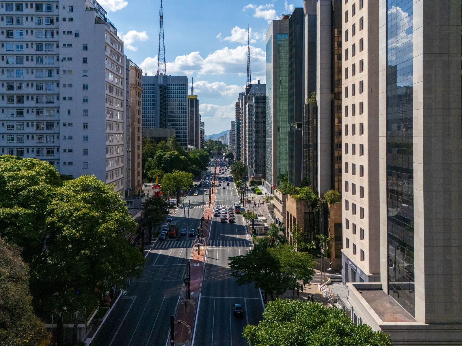 Avenida Paulista seen from above between two towers: the avenue running straight into the distance with traffic on it, street trees down both sides and glass office blocks closing it in