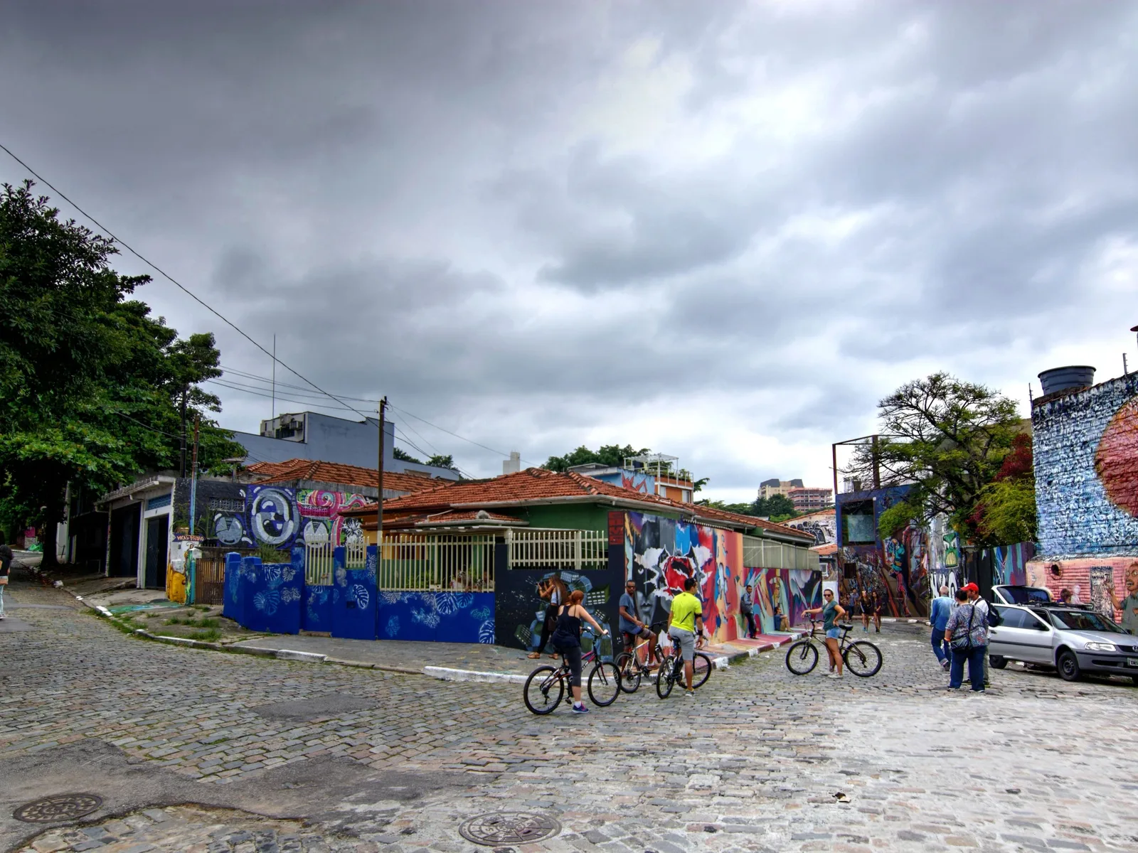 Cyclists stopped in a group on a Vila Madalena street corner in front of a long wall of bright graffiti, low houses and a red-roofed building behind them under a heavy gray sky