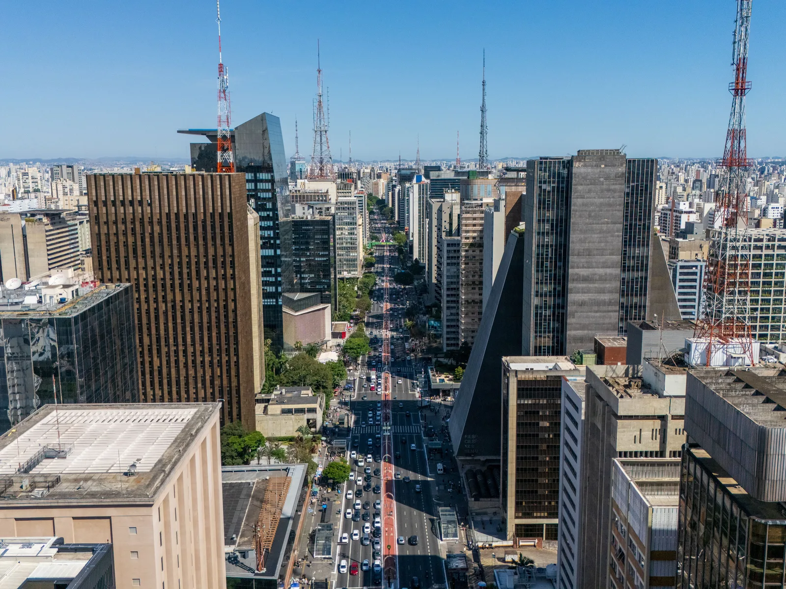 Avenida Paulista from the air along its length: office towers in glass, concrete and brown stone crowding both sides of the avenue, traffic and treetops running down the middle, and the city stretching flat to the horizon behind