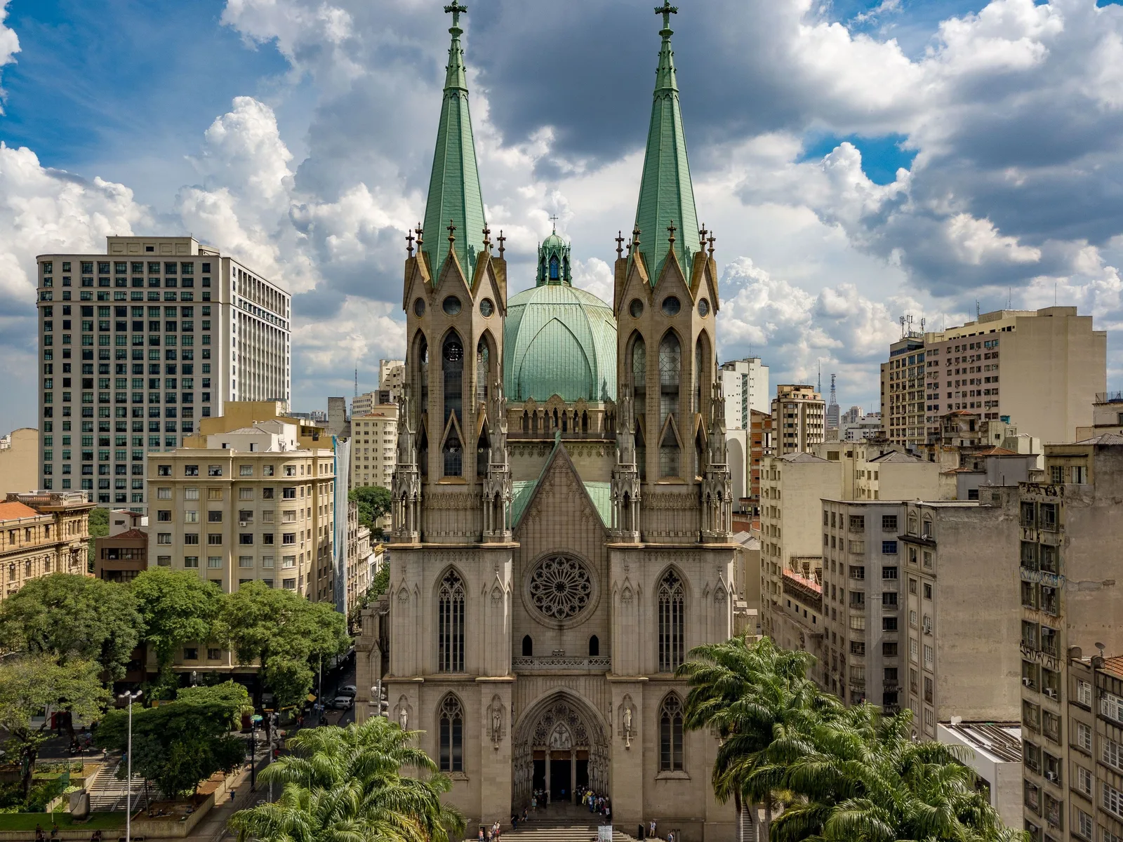The twin spires and green dome of the Catedral da Sé rising above the office blocks of central São Paulo, seen across the treetops of the square with mid-century towers packed in behind it
