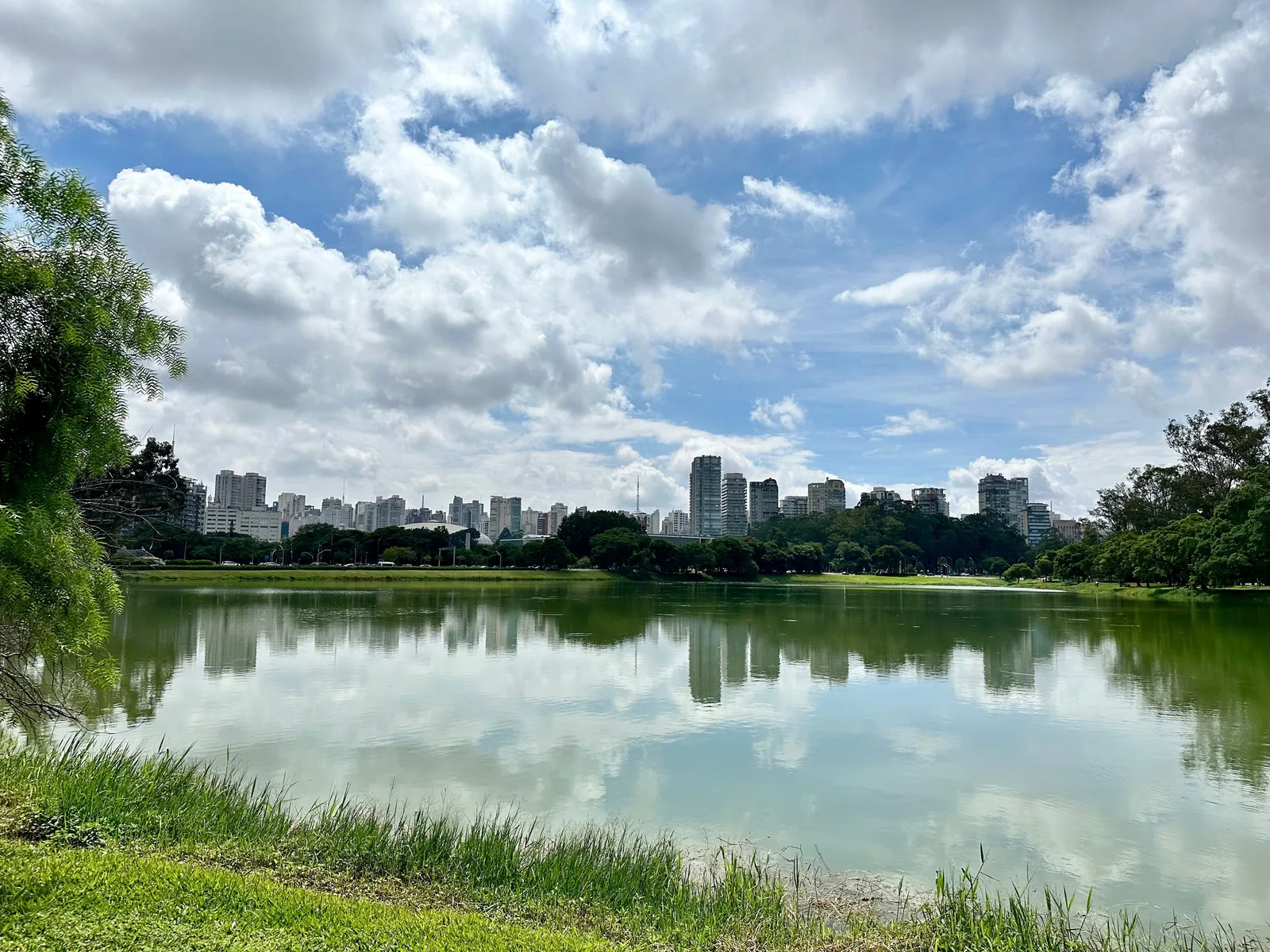 The lake at Parque Ibirapuera under a bright sky with piled white cloud, mown grass and trees along the near bank, and the low line of São Paulo’s apartment towers along the far side of the water