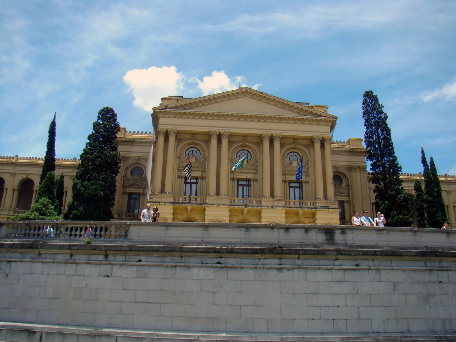 The Museu do Ipiranga seen head-on from the foot of its garden: a long pale yellow neoclassical palace with a columned central portico and flags hung from the balcony, dark cypresses standing either side of it, visitors along the balustrade above, and the blank stone face of the terrace wall filling the foreground