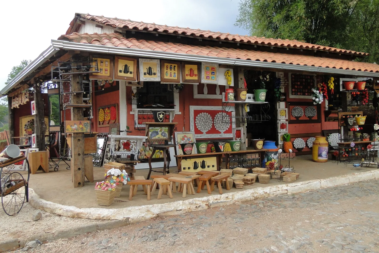 The open front of a craft shop in Bichinho under a tiled roof, its red walls hung with painted panels and mirrors and stools and carved pieces set out along the road in front