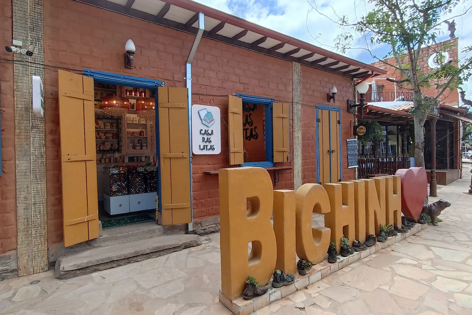 Large wooden letters spelling BICHINHO standing on the pavement outside a red-brick craft shop with an open blue-framed doorway, on the village’s single street
