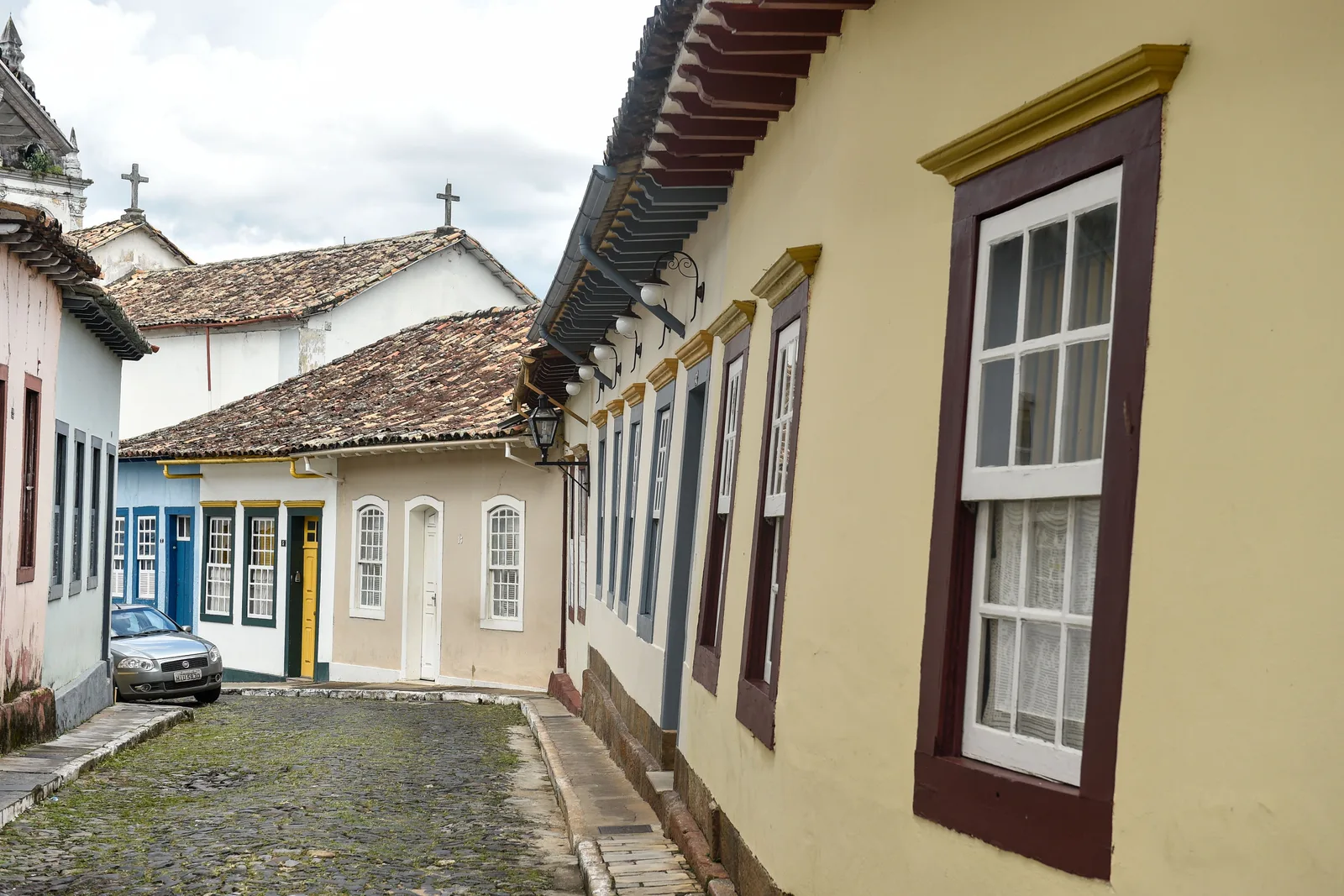 A cobbled street in São João del-Rei’s historic center: a pale yellow house with tall shuttered windows filling one side of the frame, and a row of low pink, blue and white colonial houses with visibly sagging tile roofs along the other