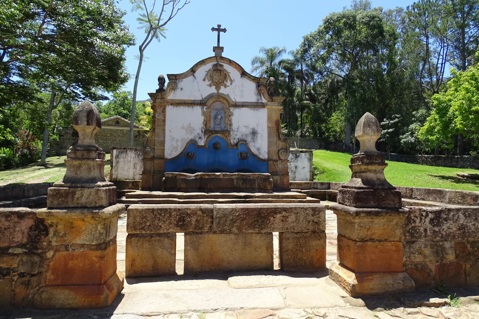 The Chafariz de São José in Tiradentes: a long stone fountain with a blue-painted niche and a painted saint above three worn basins, standing on grass with trees behind it