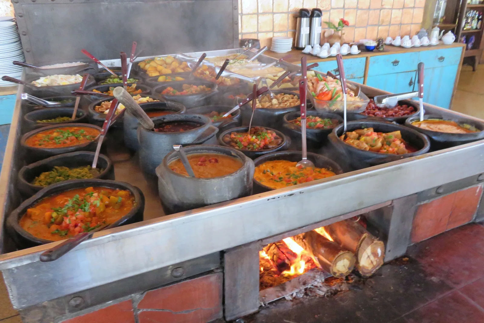 A line of dark enamel pans of stews and beans kept hot along the top of a wood-fired stove, with the fire visible burning in the open hearth underneath