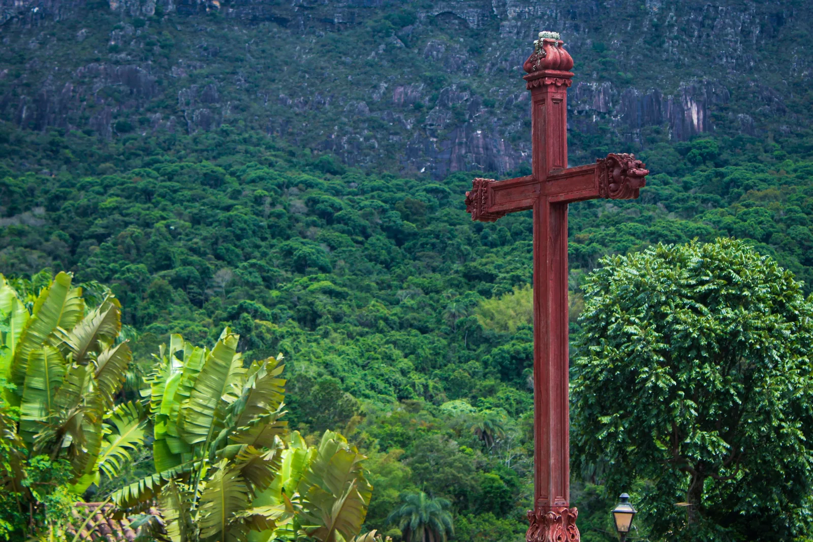 A tall red-painted timber cross standing against a solid wall of green forest, with the gray rock of the Serra de São José showing through the trees behind it