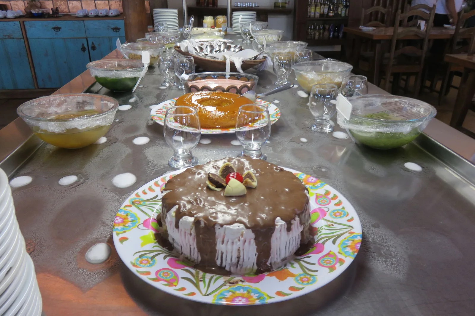 A long table set with home-made desserts: a chocolate-topped cake on a painted plate in front, glass bowls of pale puddings and compotes ranged behind it and a tiered stand at the far end