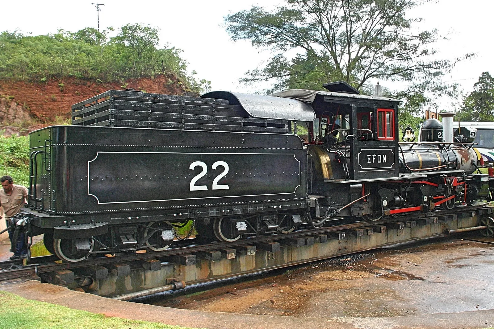 Steam locomotive number 22 standing on the turntable at the São João del-Rei works in the rain, its tender lettered with the number and the EFOM initials, with a red earth bank and trees behind