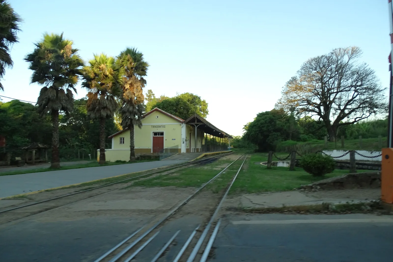 The single track running away from the low station building at Tiradentes in late afternoon light, with palms and big trees along the line and no train at the platform