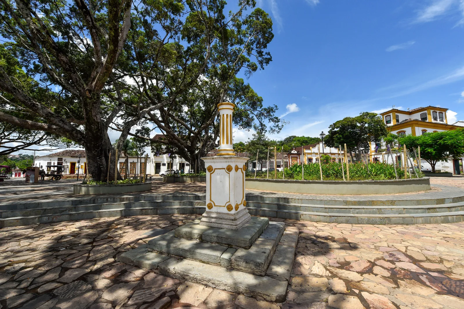 The Largo das Forras in Tiradentes: a stone monument on a stepped plinth in the middle of the paved square, under the spreading canopy of a very large tree, with white colonial buildings along the far side
