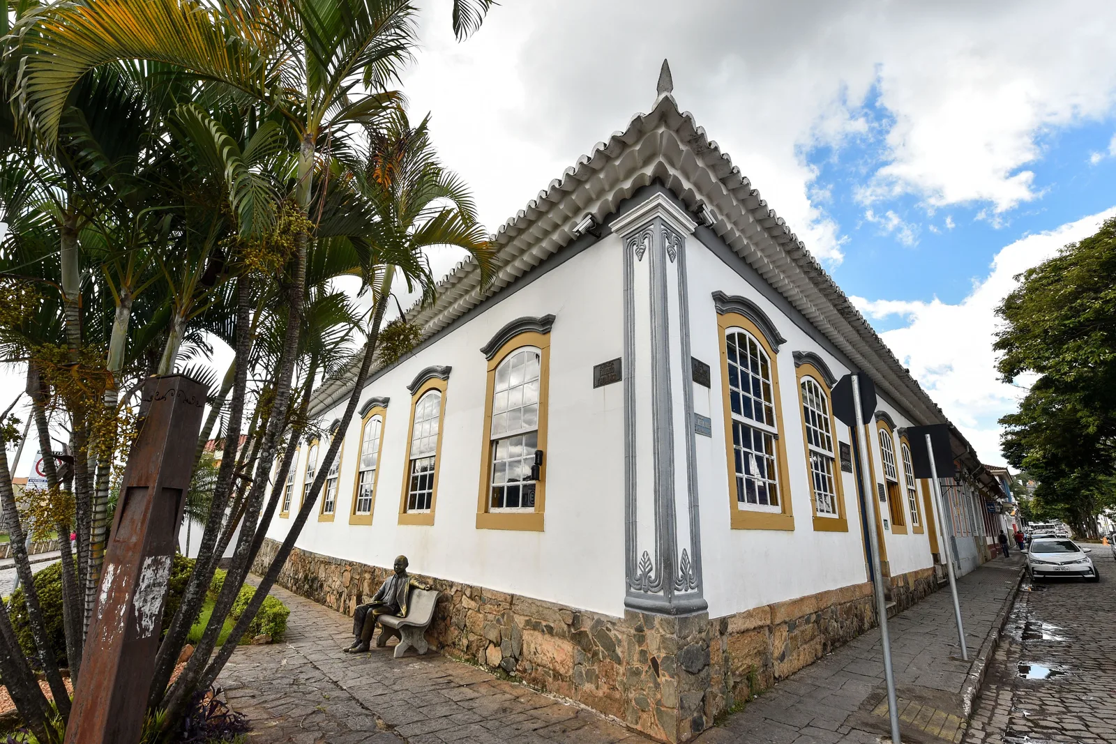 The Memorial Tancredo Neves in São João del-Rei: a low white colonial building with yellow-framed arched windows and gray stone trim on a cobbled corner, tall palms along the pavement and a bronze statue of a seated man on a bench at the foot of its wall