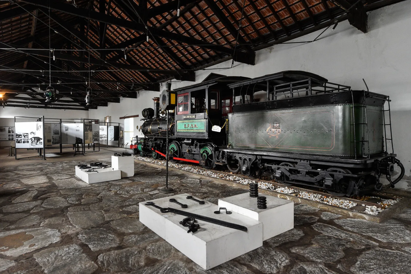 A black steam locomotive and tender standing on a stone floor inside the São João del-Rei railway museum shed, exhibition cases and display panels arranged around it under an open timber roof