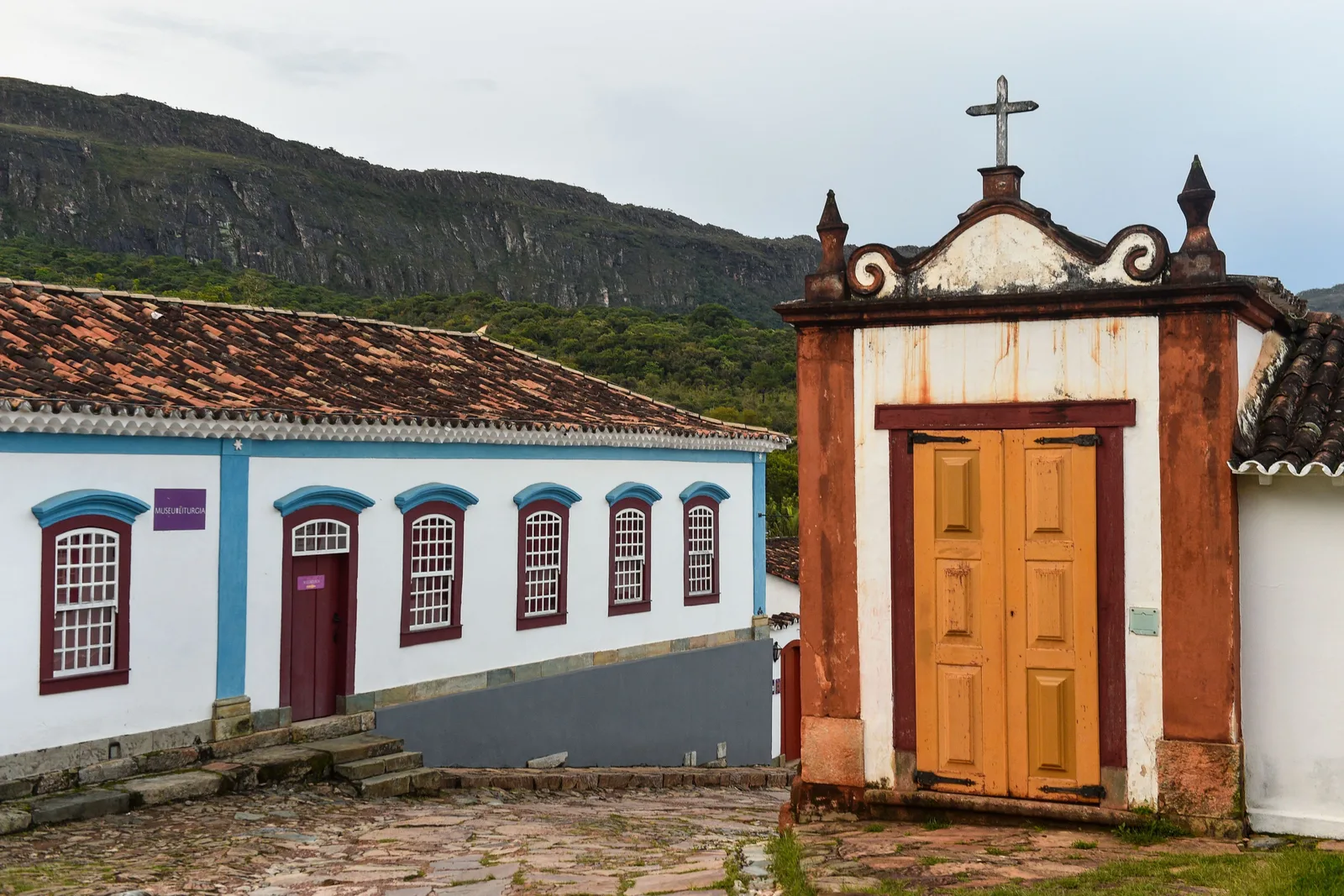 The Museu da Liturgia in Tiradentes: a long white colonial building with blue-and-red window surrounds and a small sign beside its door, on a cobbled slope next to a whitewashed gateway with yellow timber doors and a stone cross on its gable, the rock wall of the Serra de São José rising behind