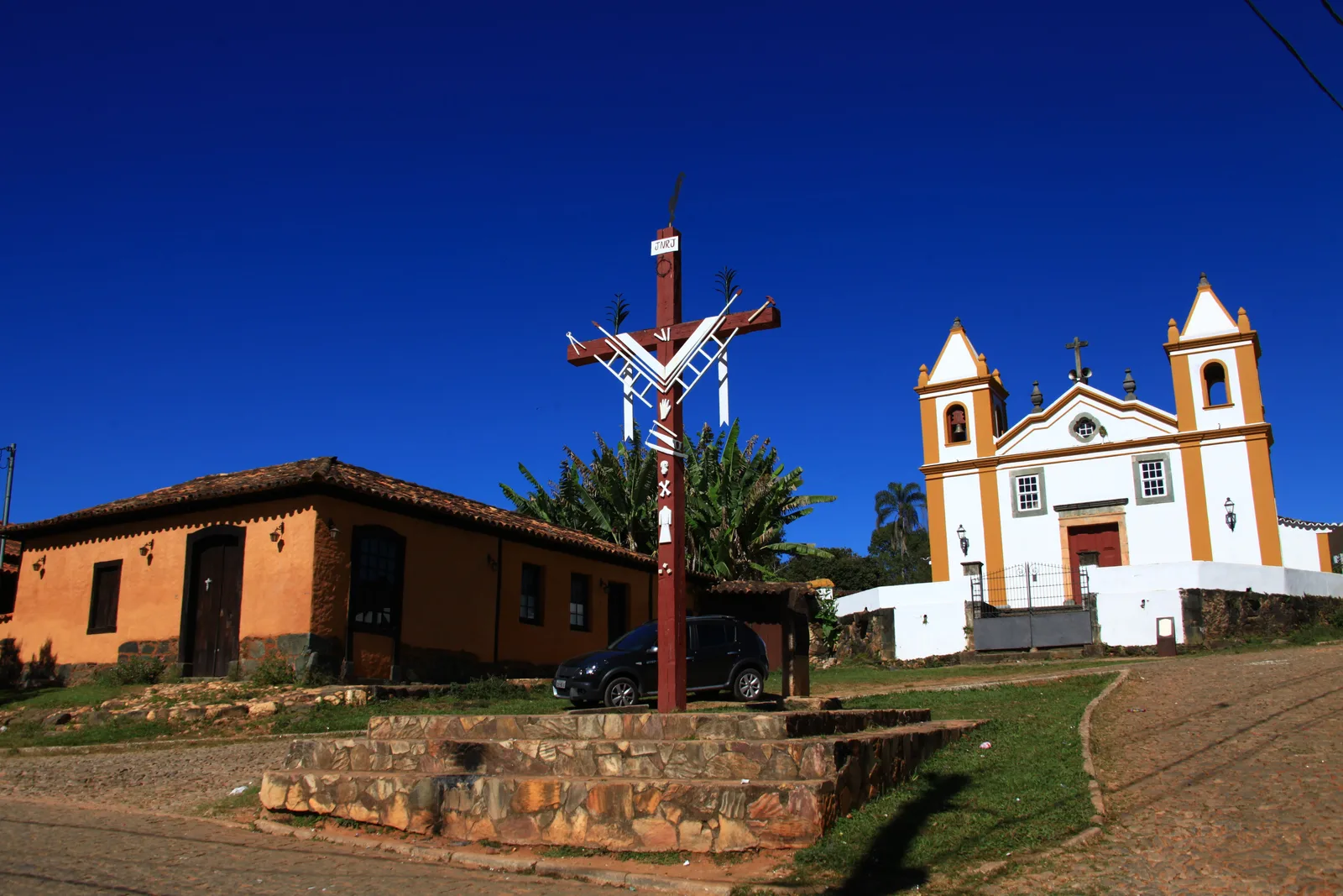 The white eighteenth-century church of Nossa Senhora da Penha at Bichinho with its twin bell towers, a tall painted timber cruzeiro on a stone base in front of it and an ochre colonial house alongside