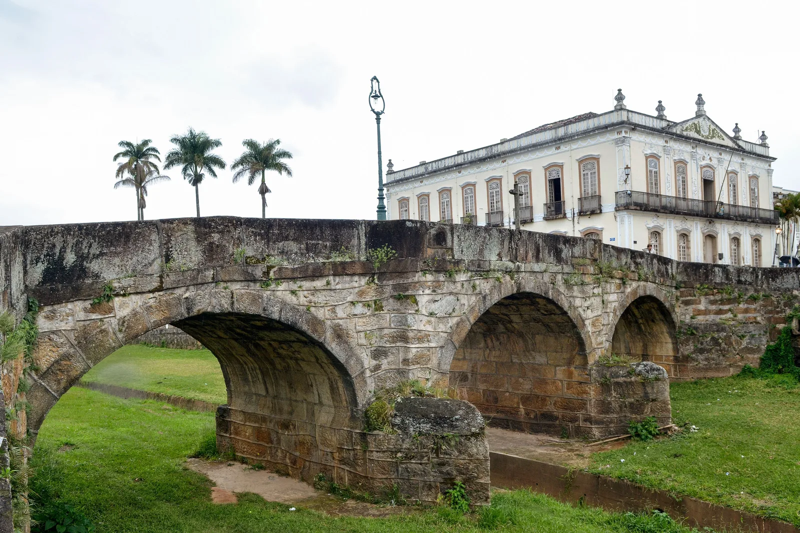 The Ponte da Cadeia in São João del-Rei, a low three-arched stone bridge over a grassy channel, with a long white colonial building and a row of palms on the bank behind it