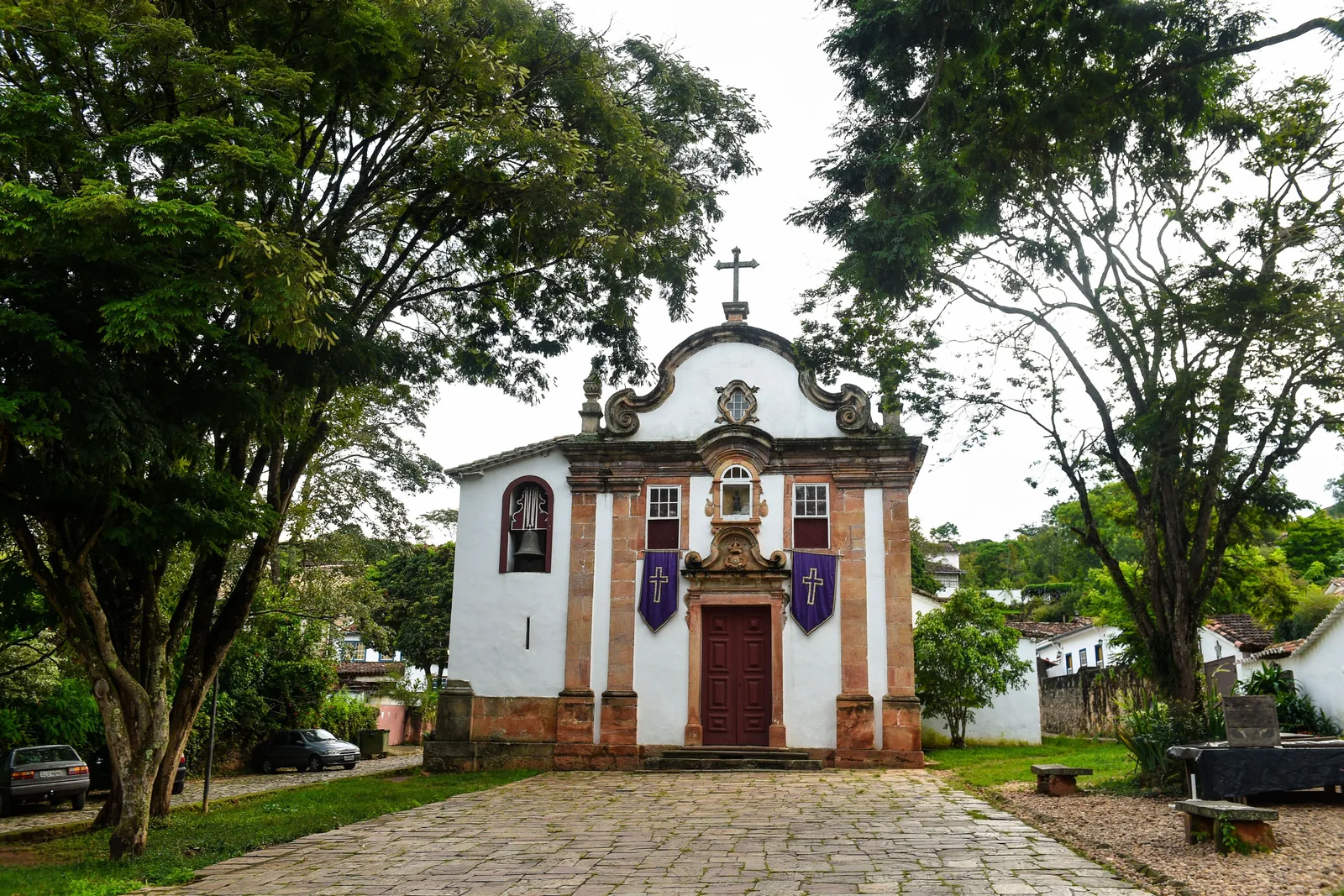 The white baroque facade of the Igreja de Nossa Senhora do Rosário dos Pretos in Tiradentes at the end of its cobbled forecourt, a cross on the scrolled gable and two purple Lenten banners hung either side of the dark red door, big trees framing it on both sides