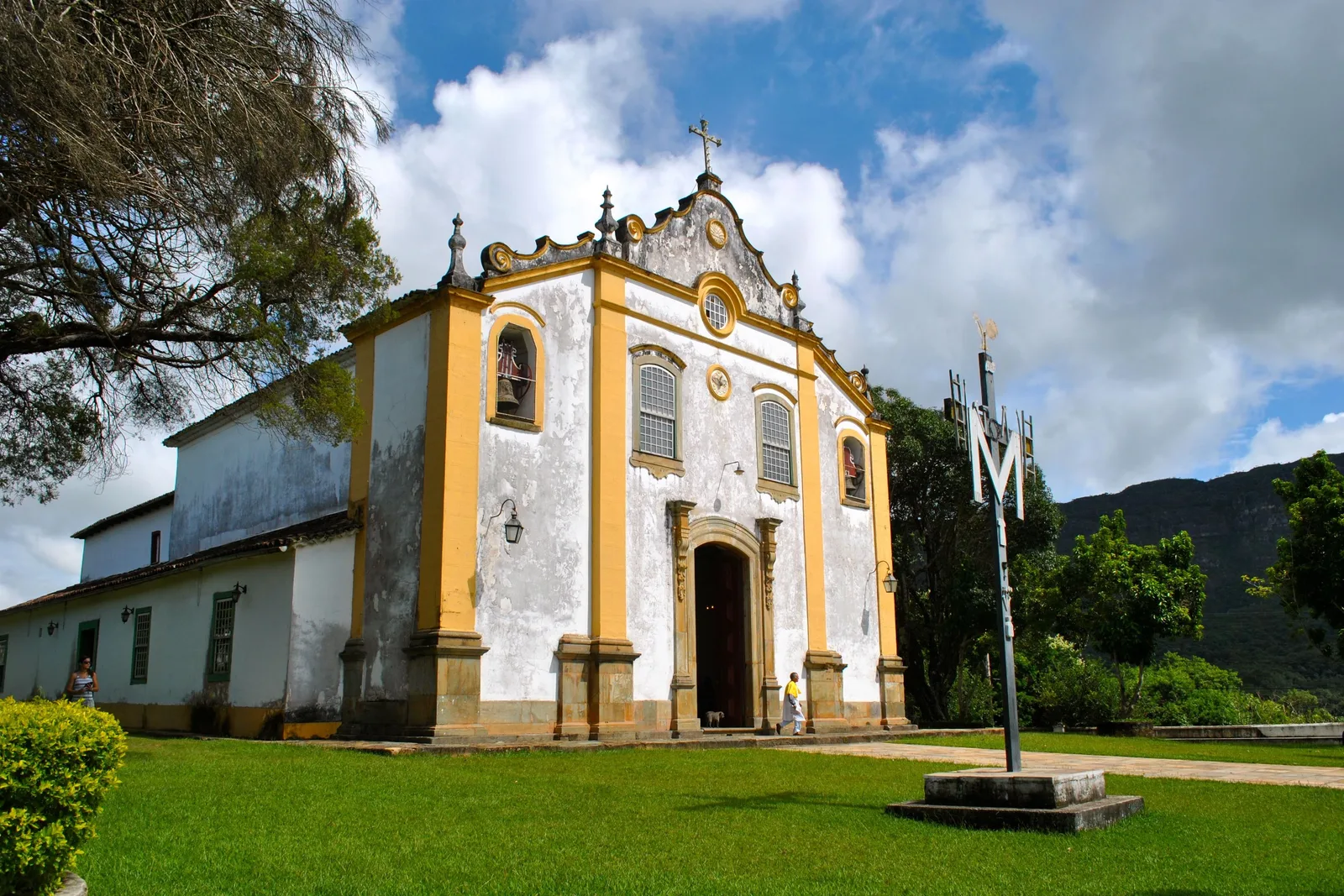 The Santuário da Santíssima Trindade on its lawn at the edge of Tiradentes, a white church with yellow-trimmed pilasters and a single central tower, with wooded hills rising behind it