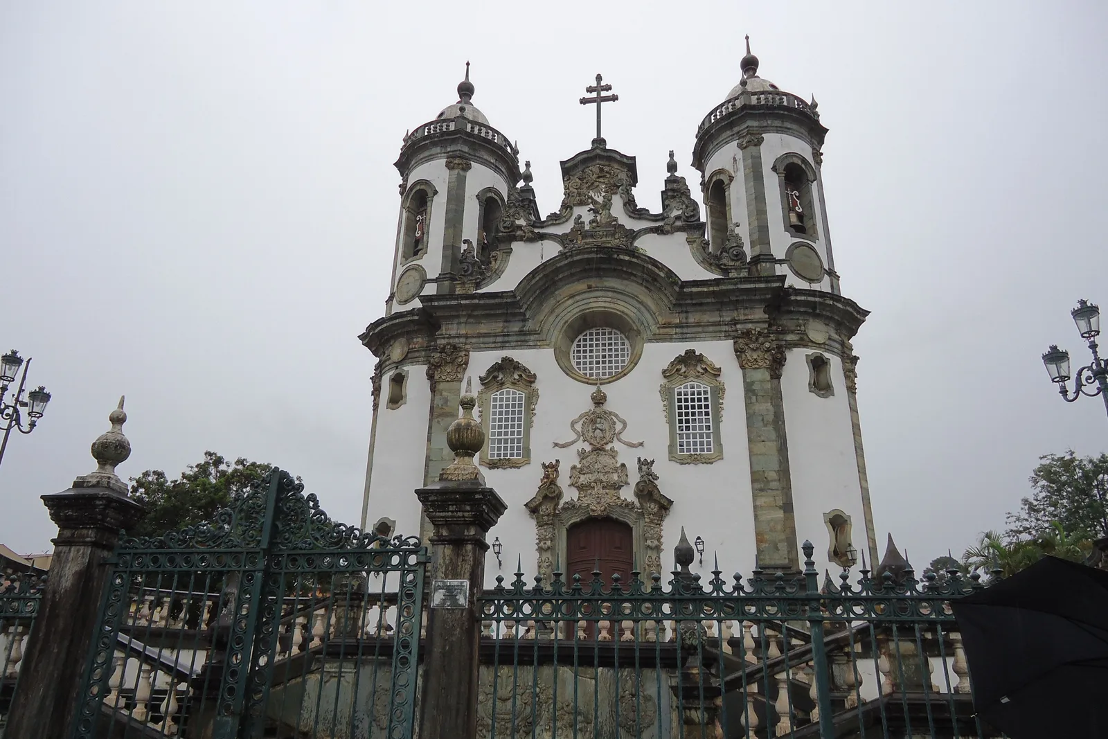 The Igreja de São Francisco de Assis in São João del-Rei behind its iron railings on an overcast day, the pale curved facade set between two rounded bell towers with a stone cross in the forecourt