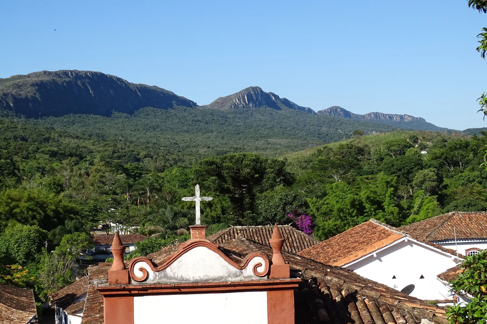 The cross on a church gable in the foreground with red-tiled rooftops below it, looking across a wooded valley to the long green ridge of the Serra de São José