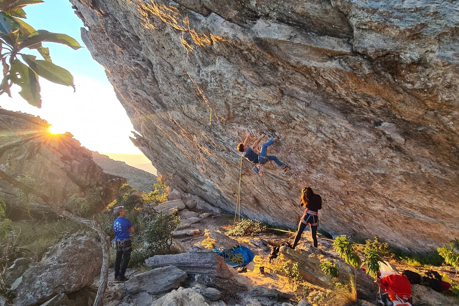 Climbers under a huge overhanging rock face on the Serra do Lenheiro at sunrise, one of them on the wall on a rope while two others stand on the boulders below with kit spread around them
