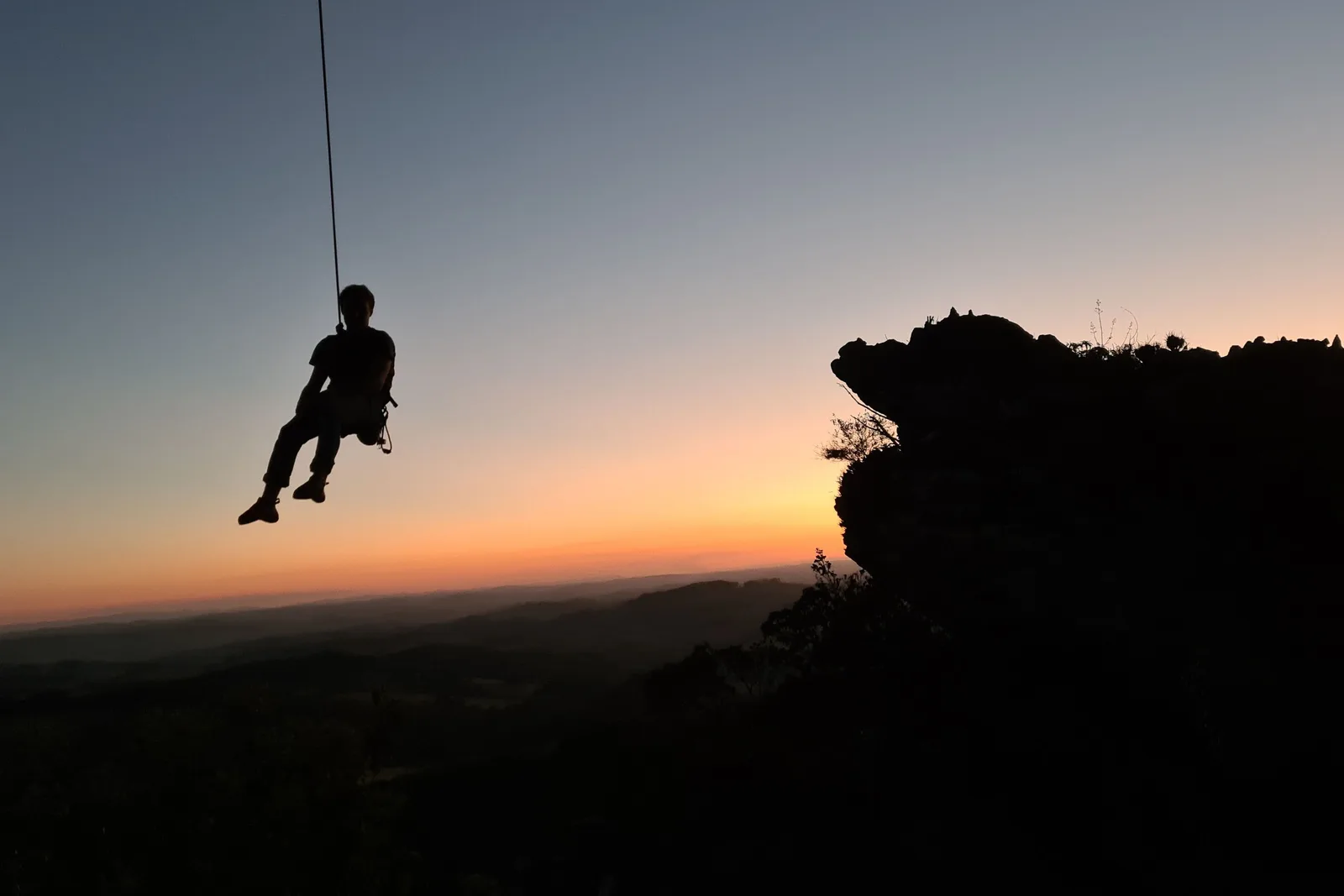 A person silhouetted on a rope part-way down a rock face at sunset, the dark shoulder of the ridge to one side and the valley haze glowing orange behind