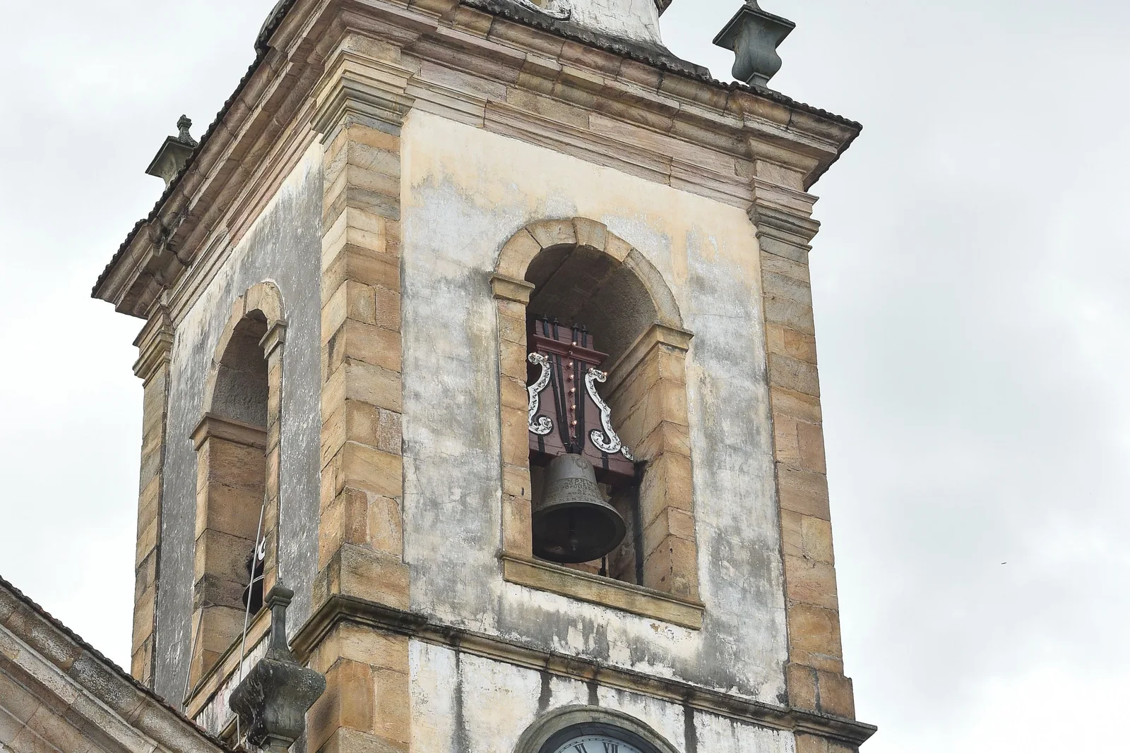 The weathered stone bell tower of the Pilar cathedral in São João del-Rei against a pale sky, one large bronze bell hanging in the open arch