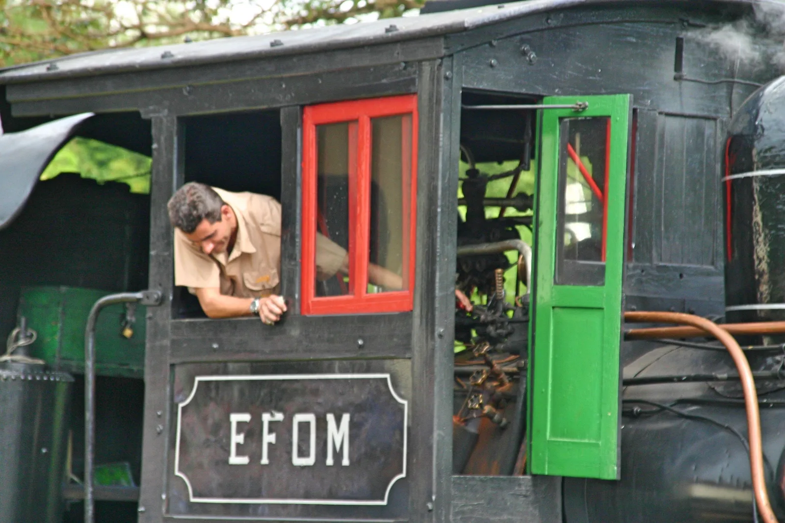 A driver leaning out of the green-and-red doorway of a steam locomotive cab, the enamel plate below him reading EFOM for the Estrada de Ferro Oeste de Minas
