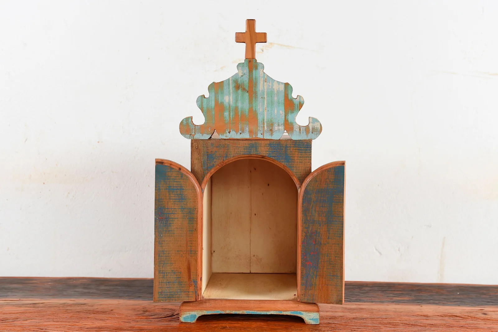 An empty wooden oratory shrine with its two doors open, painted in worn blue and terracotta and topped with a small cross, standing on a plain table