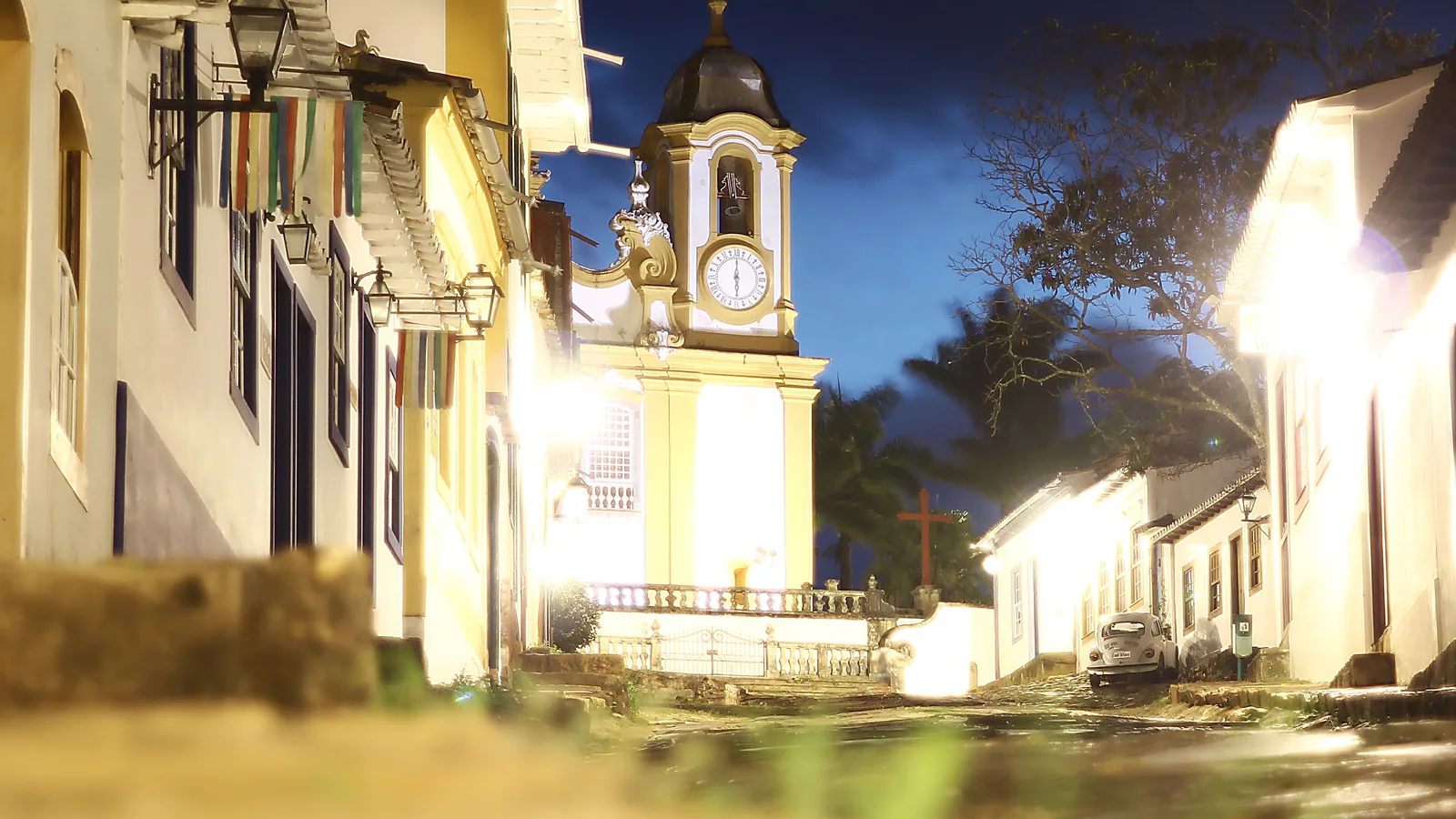 The floodlit tower of the Matriz de Santo Antônio at the top of a cobbled Tiradentes street at blue hour, its clock face and bell lit gold, lamps burning along the colonial houses on both sides and an old Volkswagen Beetle parked below