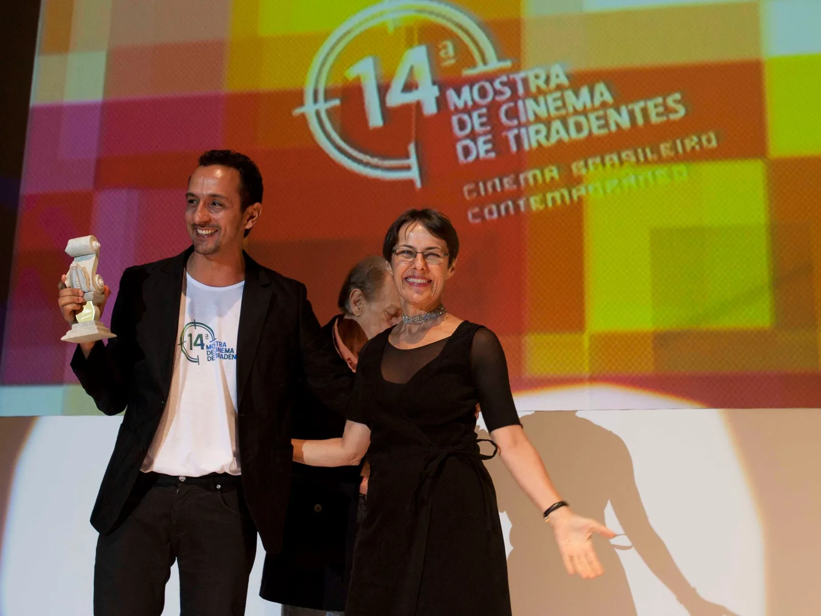 A man and a woman on stage in front of the orange backdrop of the 14th Mostra de Cinema de Tiradentes, he holding a small pale trophy, a third person half hidden behind them