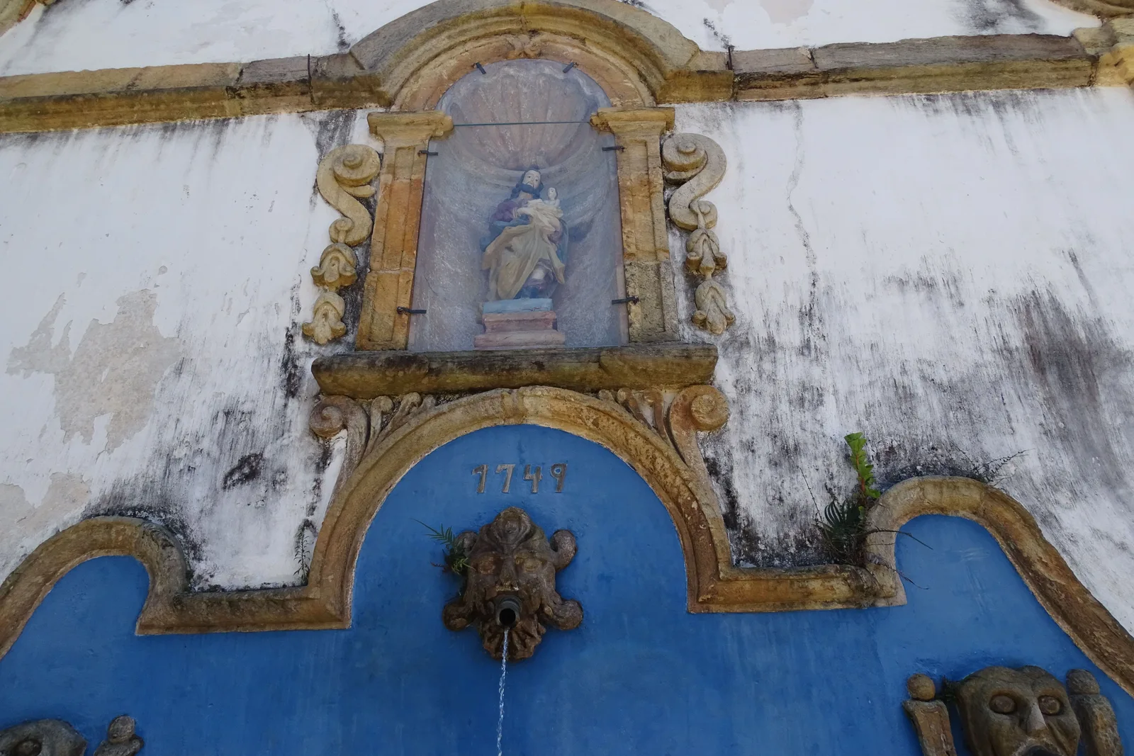 Close on the Chafariz de São José in Tiradentes: a blue-painted stone niche framing a small painted saint, with weathered bronze spouts below it