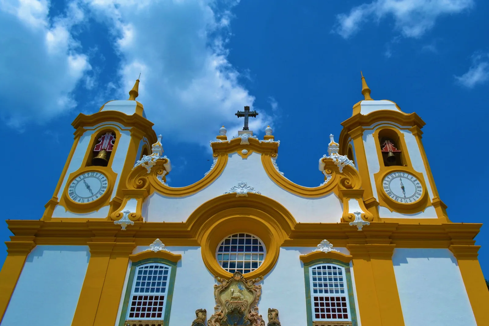 The upper facade of the Matriz de Santo Antônio in close-up: two yellow-and-white bell towers with clock faces and a carved gable between them against a deep blue sky