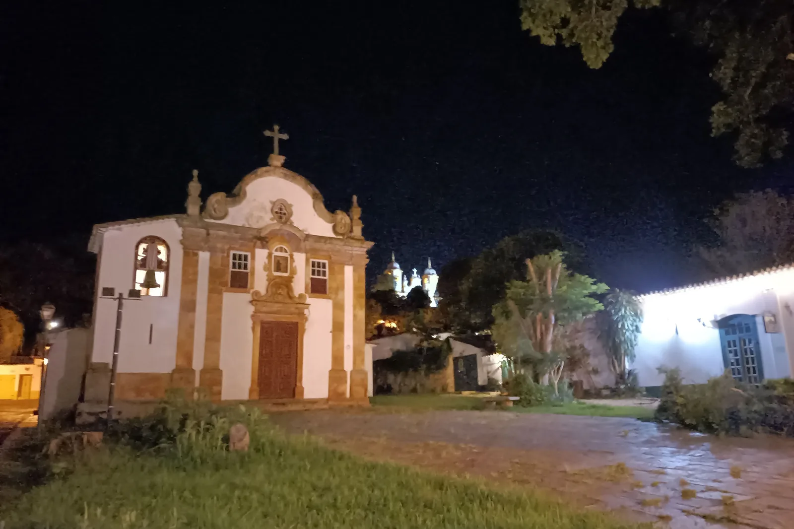 The Igreja de Nossa Senhora do Rosário dos Pretos in Tiradentes floodlit at night, its scrolled white gable and stone door surround warm against a black sky, with the lit towers of the Matriz on the hill behind it