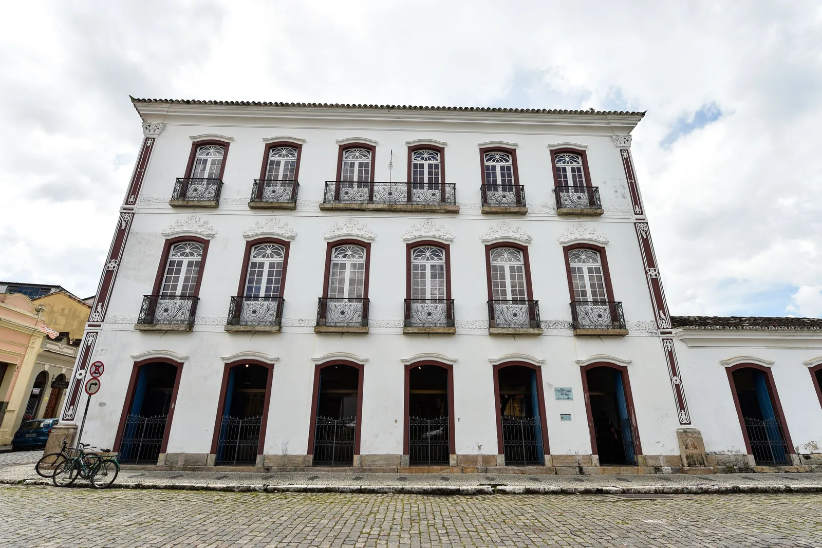 A tall white three-story colonial mansion on a cobbled square in São João del-Rei, two rows of iron balconies across its front and a line of arched doorways along the ground floor