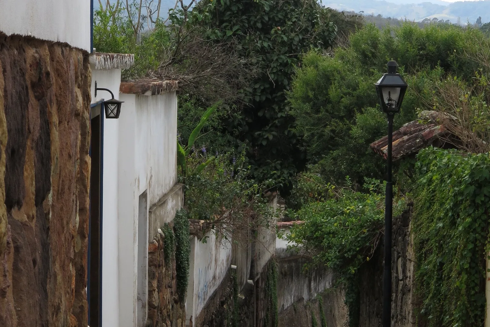 A narrow lane in Tiradentes between an old stone retaining wall and a whitewashed house, an iron street lamp on the wall and greenery spilling over the top