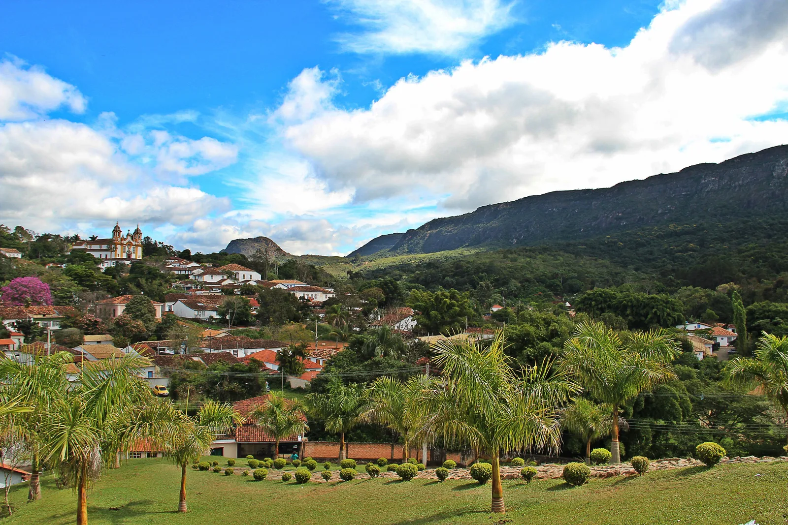Tiradentes from above the gardens on its edge: palms and dense green in front, red-tiled roofs in the middle distance and the dark wooded range beyond