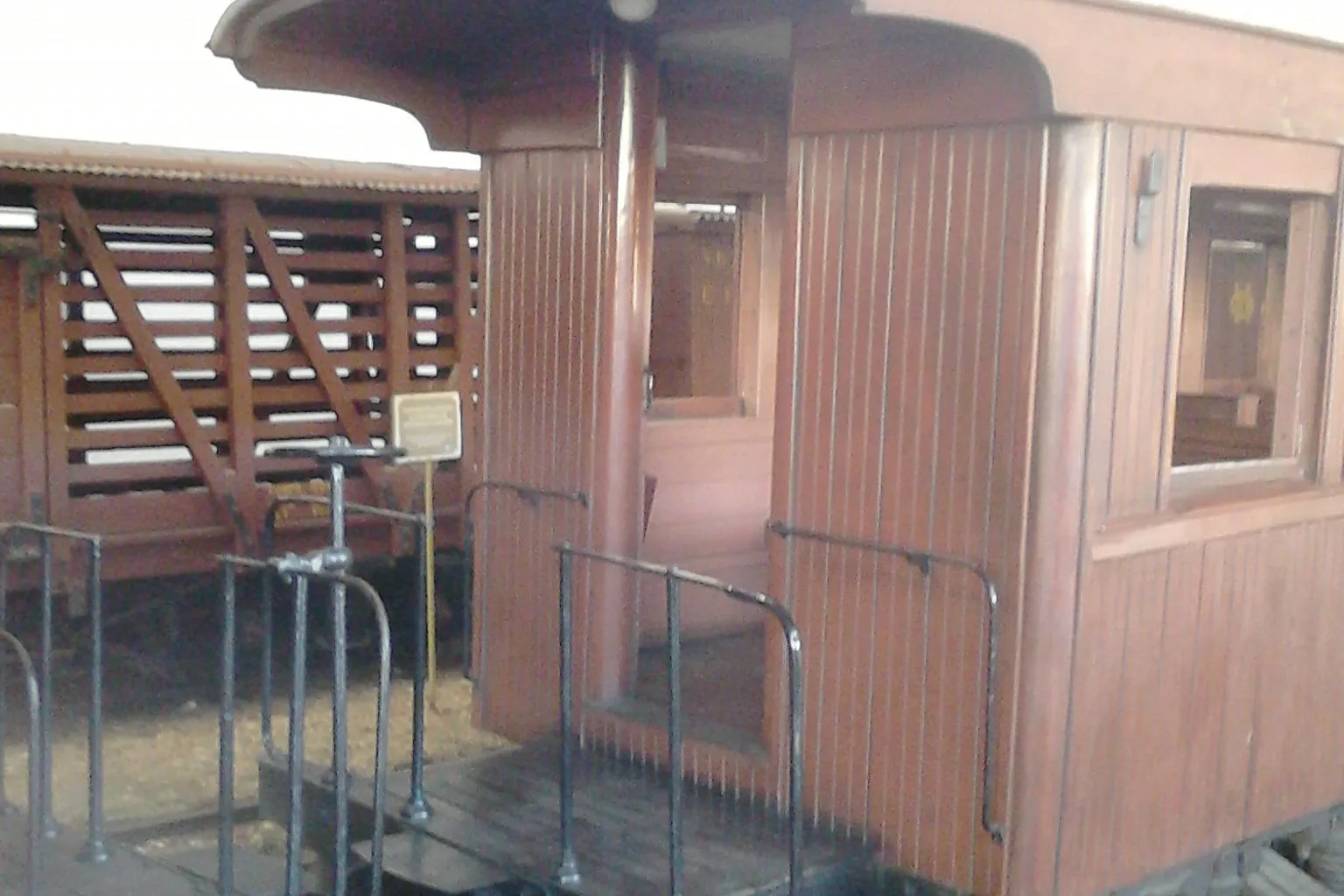 The open end platform of a wooden passenger carriage on the São João del-Rei steam railway, its slatted timber gate folded back, metal handrails on either side and a small notice board fixed to the panelling