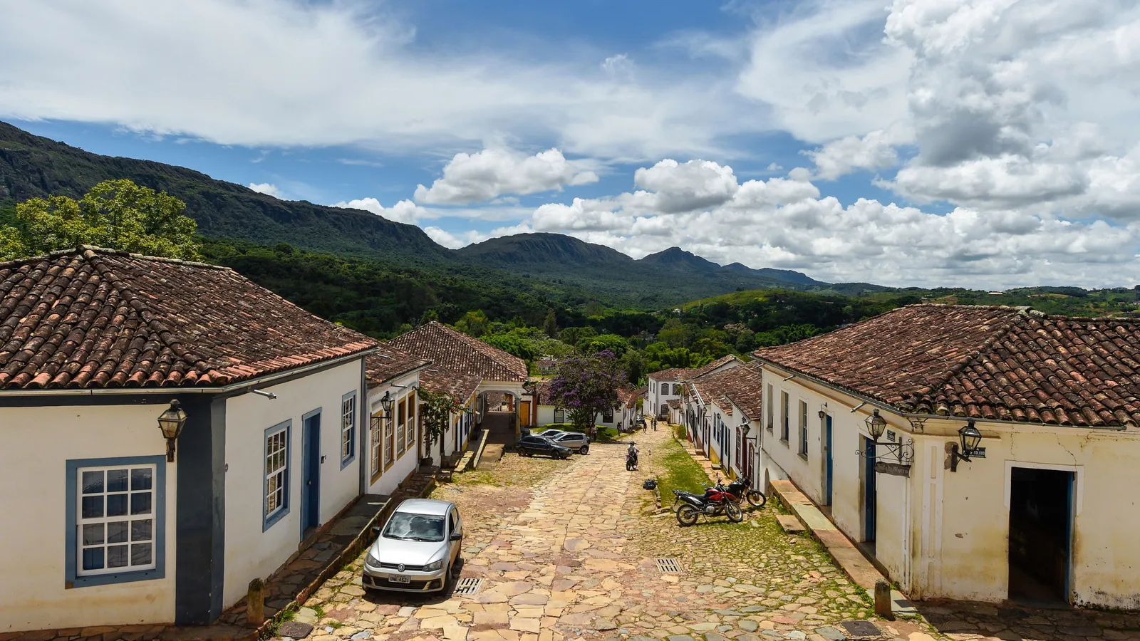 A cobbled street in Tiradentes between whitewashed colonial houses, with the wooded ridge of the Serra de São José beyond