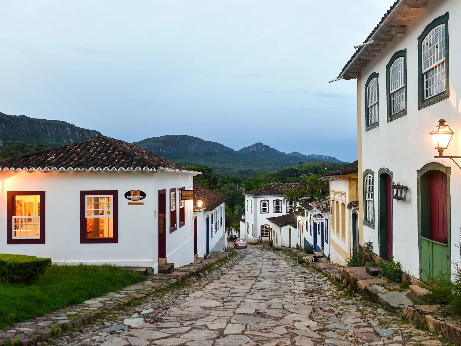 A cobbled street in Tiradentes in late afternoon light, whitewashed houses with colored doors and iron lamps running away downhill, and blue hills at the end of it
