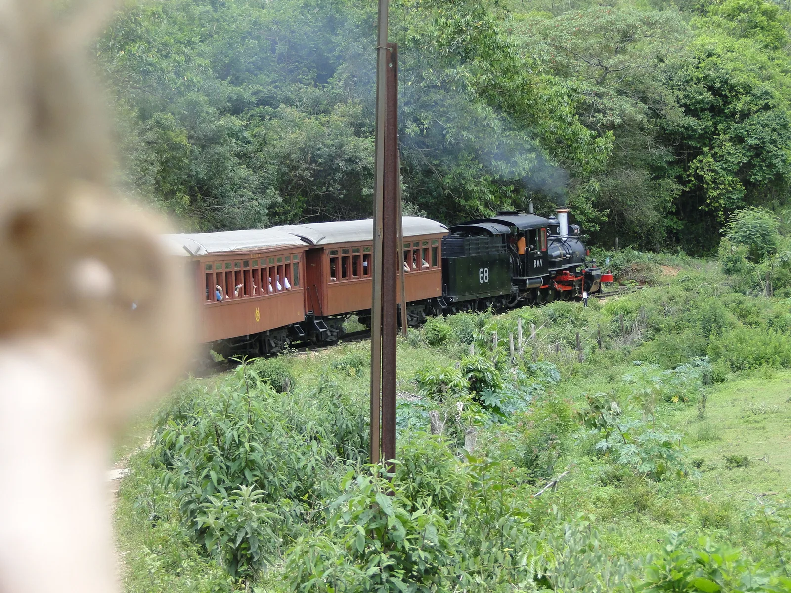 The steam train running through open green countryside between the two towns, a small black locomotive at the head of four dark red wooden carriages with wooded hills behind