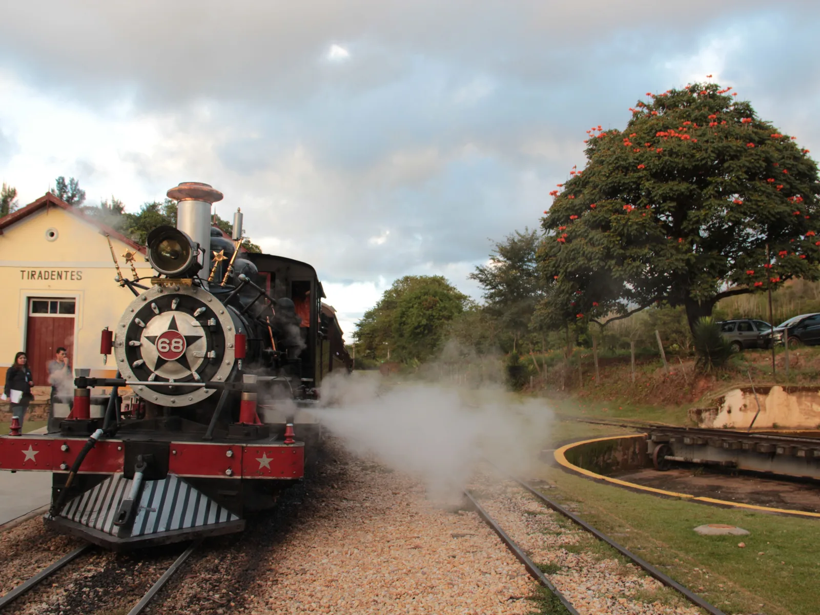 A black steam locomotive with a red-trimmed cowcatcher and a brass star on its smokebox door standing at the platform in a cloud of its own steam, the station canopy just visible at the left and green hills behind