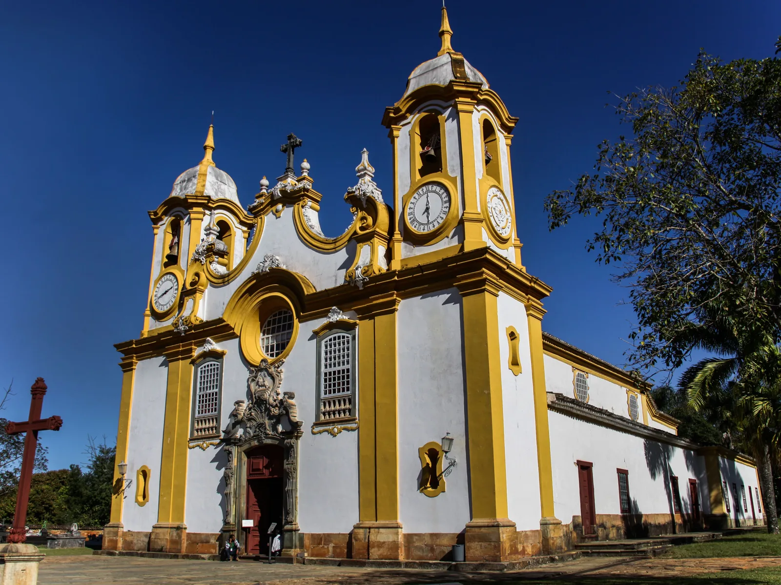 The Matriz de Santo Antônio in Tiradentes seen straight on from the terrace below: a yellow-and-white rococo facade with two bell towers, a clock face in each, a carved central gable and a stone cross standing in the foreground